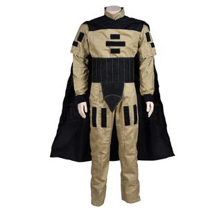 Puede incluir: Un mono beige con detalles negros y una capa negra. El mono tiene un cuello alto y un cintur&oacute;n con un dise&ntilde;o geom&eacute;trico. La capa es larga y fluida.