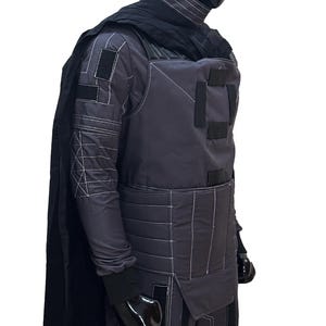 K&ouml;nnte beinhalten: Ein dunkelgraues Kost&uuml;m mit wei&szlig;en N&auml;hten, das ein gestepptes Design an den &Auml;rmeln und der Brust aufweist. Das Outfit umfasst einen schwarzen Umhang, Handschuhe und einen hochgeschlossenen Kragen. Das Kost&uuml;m wird auf einer Schaufensterpuppe pr&auml;sentiert.