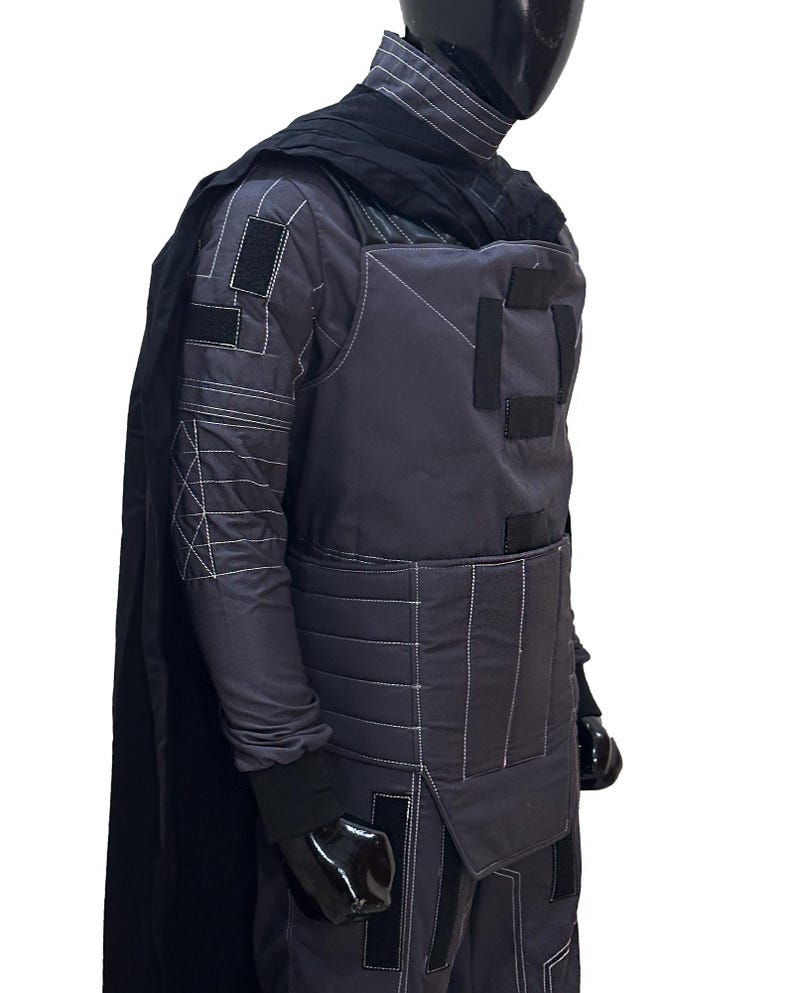 K&ouml;nnte beinhalten: Ein dunkelgraues Kost&uuml;m mit wei&szlig;en N&auml;hten, das ein gestepptes Design an den &Auml;rmeln und der Brust aufweist. Das Outfit umfasst einen schwarzen Umhang, Handschuhe und einen hochgeschlossenen Kragen. Das Kost&uuml;m wird auf einer Schaufensterpuppe pr&auml;sentiert.
