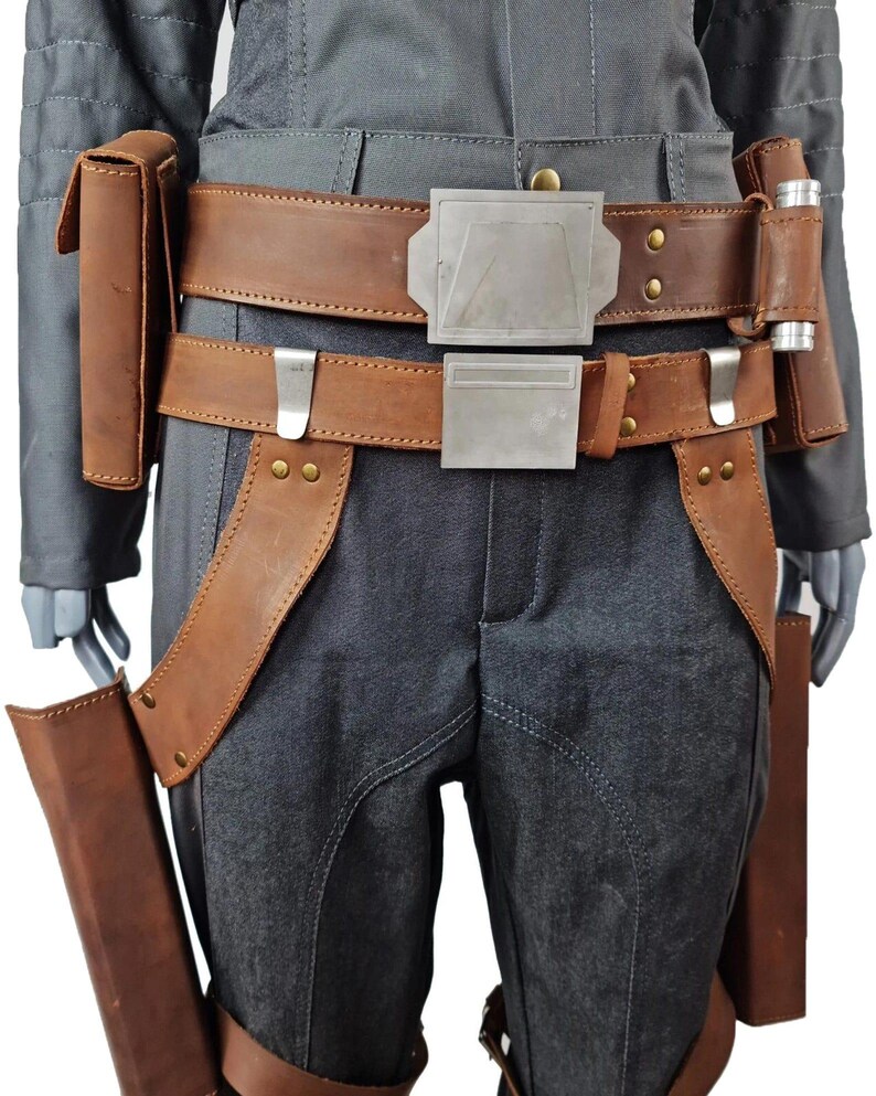 May include: A costume detail showing a grey jumpsuit with brown leather elements. It features a double belt with silver buckles, pouches, and leg holsters. The leather has a worn appearance, and the jumpsuit has visible stitching details.