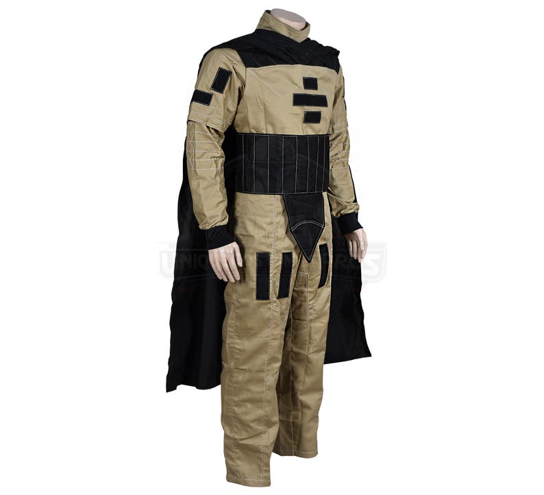 Puede incluir: Un mono beige con detalles negros y una capa negra. El mono tiene un cintur&oacute;n negro y formas geom&eacute;tricas negras en el pecho y las piernas.