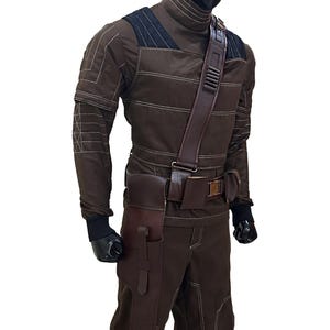 Pu&ograve; includere: Un costume marrone con cuciture bianche, con un top a collo alto, pantaloni e una cintura multiuso con tasche. Il costume include una tracolla e guanti neri. Il manichino &egrave; su sfondo bianco.