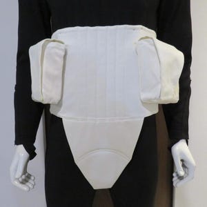 May include: A white, padded garment with a structured design, featuring a wide waistband and attached side pockets. The garment is worn over black clothing, suggesting a protective or supportive function. The design includes vertical stitching.
