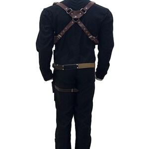 May include: Brown leather harness with adjustable straps and metal hardware. The harness is worn over a black long-sleeve shirt and pants. A matching belt and leg strap are also visible. The outfit is displayed on a mannequin.