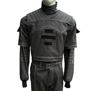 May include: A dark gray jumpsuit with a high collar and white stitching. The outfit features padded shoulders, short sleeves, and long sleeves underneath. Two black rectangular patches are on the chest. The pants have a belt and the sleeves have black cuffs.