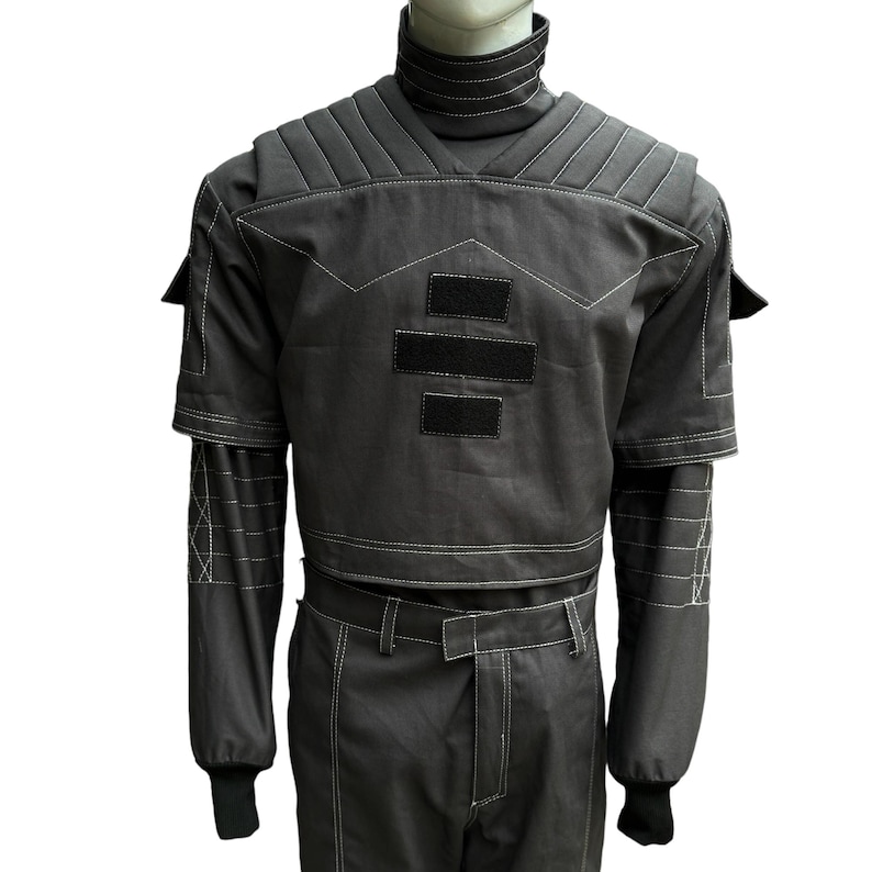 May include: A dark gray jumpsuit with a high collar and white stitching. The outfit features padded shoulders, short sleeves, and long sleeves underneath. Two black rectangular patches are on the chest. The pants have a belt and the sleeves have black cuffs.