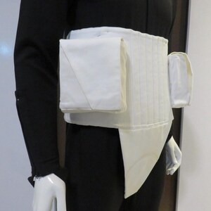 Puede incluir: Un cintur&oacute;n utilitario blanco con m&uacute;ltiples bolsillos y un dise&ntilde;o acolchado. El cintur&oacute;n tiene un bolsillo rectangular en el lado izquierdo y un bolsillo m&aacute;s peque&ntilde;o y redondeado en el lado derecho. El cintur&oacute;n se usa sobre un atuendo negro.