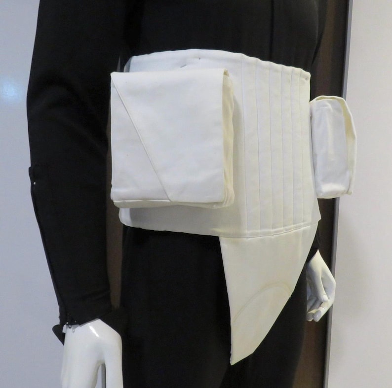 Puede incluir: Un cintur&oacute;n utilitario blanco con m&uacute;ltiples bolsillos y un dise&ntilde;o acolchado. El cintur&oacute;n tiene un bolsillo rectangular en el lado izquierdo y un bolsillo m&aacute;s peque&ntilde;o y redondeado en el lado derecho. El cintur&oacute;n se usa sobre un atuendo negro.