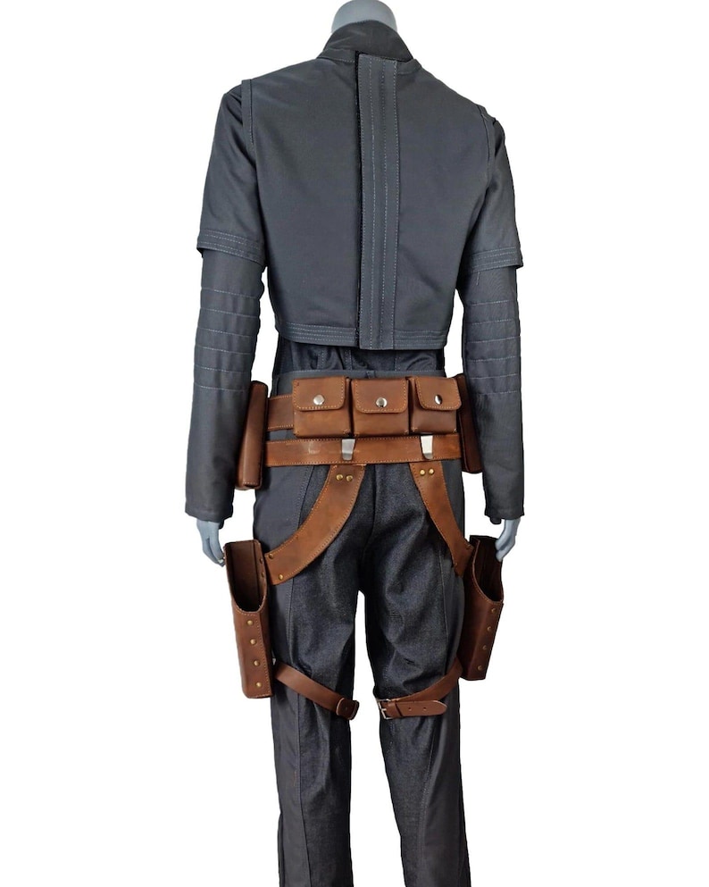 May include: A grey jumpsuit with a matching jacket and brown leather accessories. The outfit includes a belt with pouches, suspenders, and holsters. The jacket has long sleeves and a high collar.