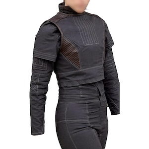 May include: A dark gray cropped jacket with brown leather accents and white stitching. The jacket has a high collar and long sleeves with quilted detailing. The matching pants are also dark gray. The outfit is a modern take on a futuristic style.