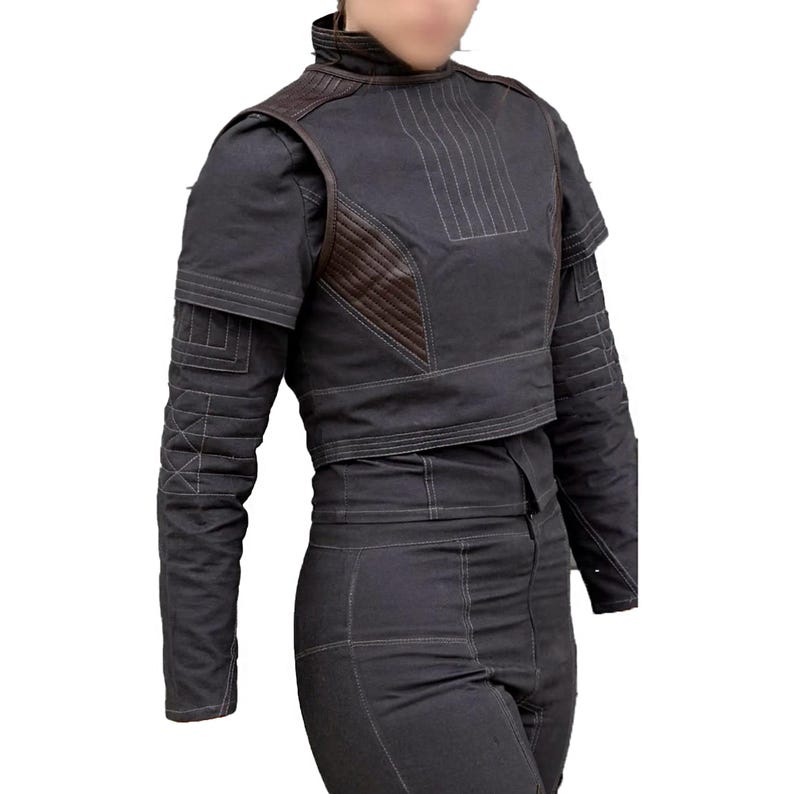 May include: A dark gray cropped jacket with brown leather accents and white stitching. The jacket has a high collar and long sleeves with quilted detailing. The matching pants are also dark gray. The outfit is a modern take on a futuristic style.