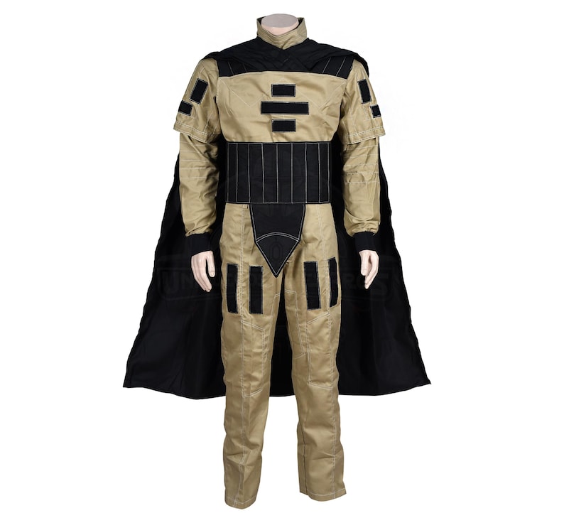 Puede incluir: Un mono beige con detalles negros y una capa negra. El mono tiene un cuello alto y un cintur&oacute;n con un dise&ntilde;o geom&eacute;trico. La capa es larga y fluida.