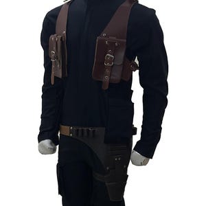 May include: Brown leather shoulder holster with two pouches and a belt with ammunition holders. The holster is worn over a black long-sleeve shirt and pants. The design includes buckles and straps for a secure fit.
