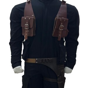 May include: A brown leather tactical harness with two shoulder pouches and a belt with a holster. The harness is worn over a black long-sleeve shirt and pants. The pouches and holster have silver buckles.