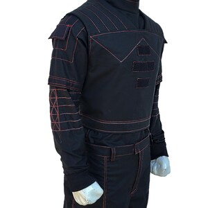 K&ouml;nnte beinhalten: Schwarzer Overall mit roten N&auml;hten. Das Outfit hat einen hohen Kragen, Schulterdetails und Taschen. Rote N&auml;hte bilden geometrische Muster auf den &Auml;rmeln und der Brust. Die Hose hat einen G&uuml;rtel und Taschen. Der Overall wird auf einer Schaufensterpuppe pr&auml;sentiert.