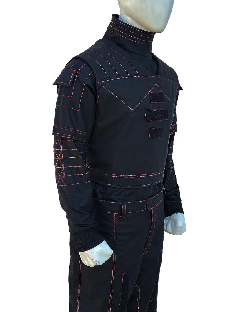K&ouml;nnte beinhalten: Schwarzer Overall mit roten N&auml;hten. Das Outfit hat einen hohen Kragen, Schulterdetails und Taschen. Rote N&auml;hte bilden geometrische Muster auf den &Auml;rmeln und der Brust. Die Hose hat einen G&uuml;rtel und Taschen. Der Overall wird auf einer Schaufensterpuppe pr&auml;sentiert.