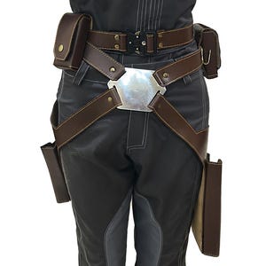 Puede incluir: Un cintur&oacute;n de cuero marr&oacute;n con una hebilla plateada y m&uacute;ltiples bolsas. El cintur&oacute;n se lleva sobre pantalones grises oscuros.