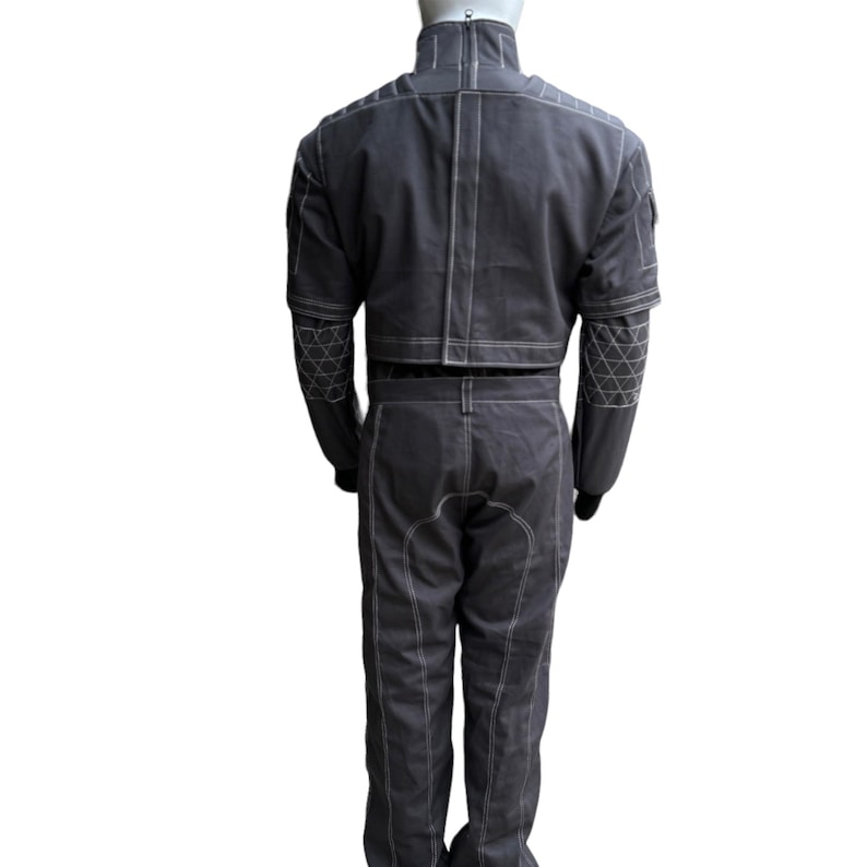 May include: A dark gray jumpsuit with white stitching and geometric patterns on the sleeves. The outfit includes a cropped jacket and matching pants. The jumpsuit is displayed on a mannequin against a white background.