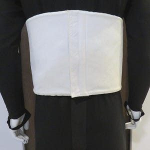 Puede incluir: Un cors&eacute; blanco rectangular usado sobre un atuendo negro. El cors&eacute; tiene un acabado liso y parece estar hecho de una tela resistente. Las mangas largas del atuendo son negras, con guantes blancos. Prenda de soporte.