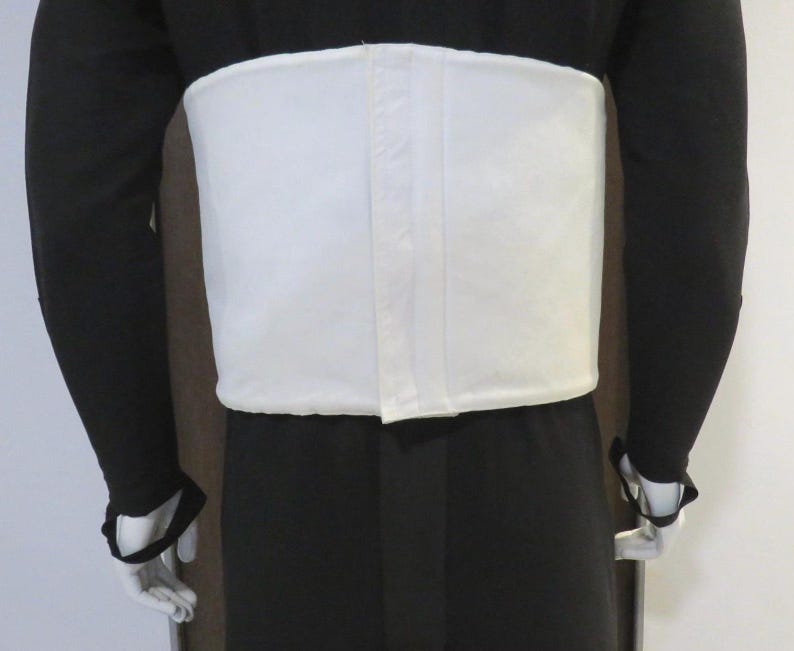 Puede incluir: Un cors&eacute; blanco rectangular usado sobre un atuendo negro. El cors&eacute; tiene un acabado liso y parece estar hecho de una tela resistente. Las mangas largas del atuendo son negras, con guantes blancos. Prenda de soporte.