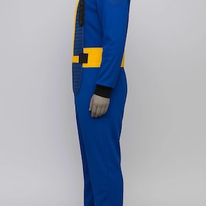 Op de afbeelding: Blauw jumpsuit met gele accenten, waaronder een gele kraag, een verticale gele streep en een gele riem. De mouwen hebben zwarte manchetten en er is een ronde patch op de schouder.