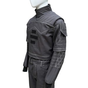 May include: A dark gray, two-piece outfit with white stitching details. The top features a high collar, shoulder padding, and three black rectangular patches. The long-sleeved jacket has a small pocket and geometric stitching on the forearm. The pants have a belt and white stitching.