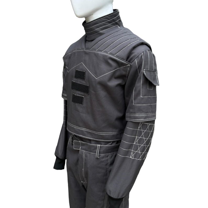 May include: A dark gray, two-piece outfit with white stitching details. The top features a high collar, shoulder padding, and three black rectangular patches. The long-sleeved jacket has a small pocket and geometric stitching on the forearm. The pants have a belt and white stitching.
