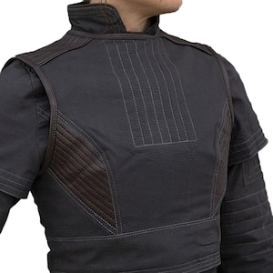 May include: A dark gray jacket with a high collar and brown leather-like accents. The jacket features vertical stitching on the front and quilted detailing on the shoulders and sleeves. The design is a blend of modern and utilitarian styles.