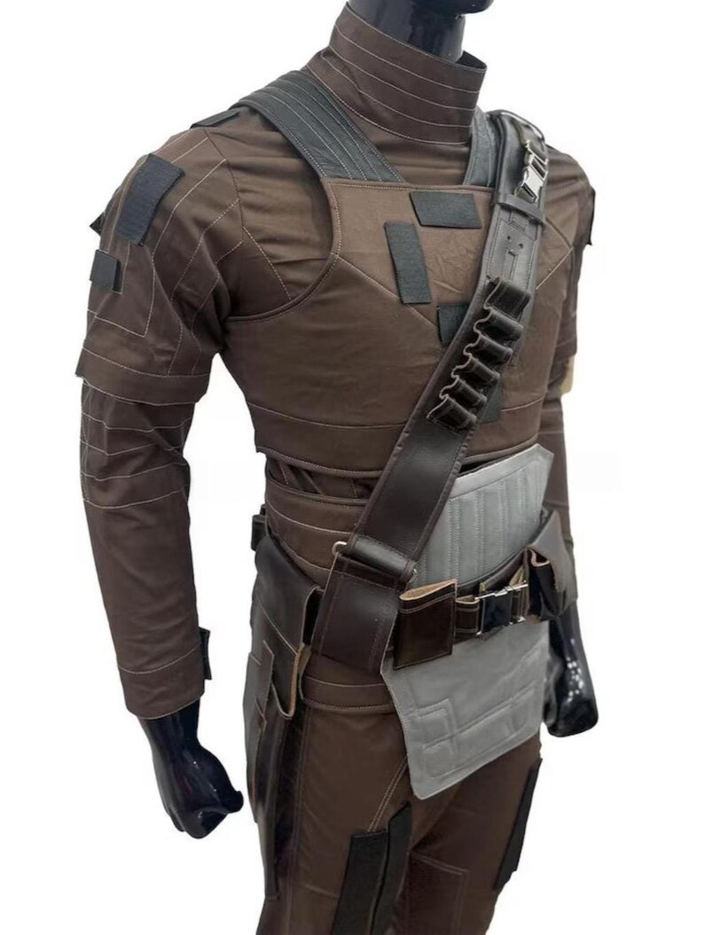 May include: A brown costume with a high collar, shoulder straps, and a utility belt. The outfit includes a chest piece, long sleeves, and a holster. The costume has black accents and a gray panel on the waist.