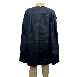 Puede incluir: Una capa negra con capucha, cuello beige y un peque&ntilde;o parche en el hombro derecho. La capa se lleva sobre una camisa y pantalones beige.