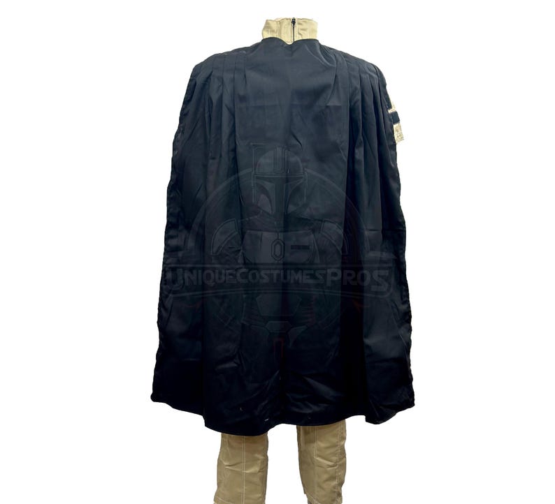 Puede incluir: Una capa negra con capucha, cuello beige y un peque&ntilde;o parche en el hombro derecho. La capa se lleva sobre una camisa y pantalones beige.