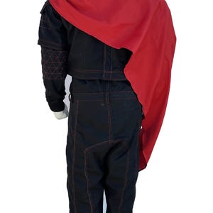 K&ouml;nnte beinhalten: Schwarzer Overall mit roten N&auml;hten und einem passenden roten Umhang. Der Overall hat einen hohen Kragen, lange &Auml;rmel und eine taillierte Silhouette. Der rote Umhang drapiert &uuml;ber den Schultern und verleiht dem Outfit einen dramatischen Flair. Das Outfit wird auf einer Schaufensterpuppe pr&auml;sentiert.