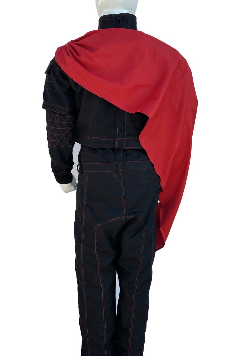 K&ouml;nnte beinhalten: Schwarzer Overall mit roten N&auml;hten und einem passenden roten Umhang. Der Overall hat einen hohen Kragen, lange &Auml;rmel und eine taillierte Silhouette. Der rote Umhang drapiert &uuml;ber den Schultern und verleiht dem Outfit einen dramatischen Flair. Das Outfit wird auf einer Schaufensterpuppe pr&auml;sentiert.