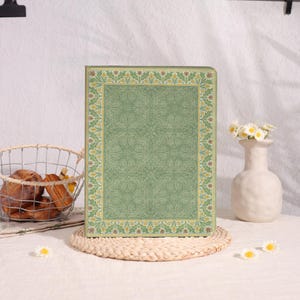May include: A rectangular, green decorative box with a floral pattern and a decorative border. The box is sitting on a woven placemat. A vase with white flowers and a basket of bread are in the background.