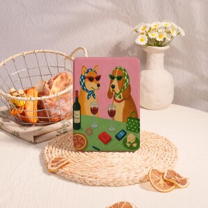 May include: A pink art print depicting two dogs in sunglasses and headscarves, drinking wine. A wine bottle, glasses, and snacks are on the table. A basket of bread and flowers are in the background.