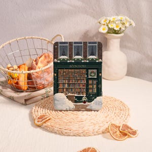 Könnte beinhalten: Dekorative Buchladen-Figur mit grüner Fassade, Obergeschoss aus Backstein und dem Wort "BOOKSTORE" über dem Eingang. Das Innere ist mit Bücherregalen gefüllt. Schnee und ein kleiner Weihnachtsbaum befinden sich vor dem Geschäft. Ein Brotkorb und eine Blumenvase sind in der Nähe.