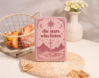 May include: A rectangular pink tin box featuring a dark red illustration of a sword, mountains, stars, and the text "the stars who listen". The tin is placed on a round woven mat, surrounded by dried citrus slices and a wire basket.