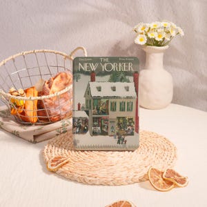 May include: A vintage copy of The New Yorker magazine, featuring a winter scene on the cover. The magazine is displayed on a woven placemat, with a basket of bread and a vase of flowers in the background. The cover shows a snow-covered building.