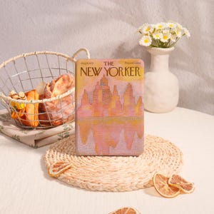 May include: A decorative clutch featuring a vintage "The New Yorker" cover illustration. The cover art depicts a cityscape in shades of pink, orange, and yellow. The clutch is displayed on a woven placemat with dried orange slices.