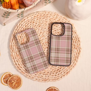 May include: Two phone cases featuring a pink and brown plaid design. One case is flat, the other upright. They rest on a woven, circular mat. The cases have a black border. The cases are for a mobile phone.