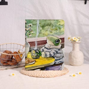 Op de afbeelding: Een rechthoekig notitieboek met een aquarelillustratie van een groene kikker in een grijze trui, zittend aan een gele tafel met een dampende mok en een croissant. De achtergrond toont een raam met groen gebladerte en een vaas bloemen.