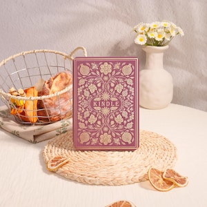 May include: A Kindle e-reader with a decorative cover featuring a floral and pomegranate design in gold on a mauve background. The e-reader is on a woven placemat, with a basket of bread and a vase of flowers in the background.