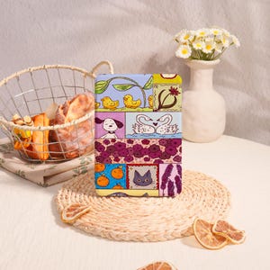 Könnte beinhalten: Ein farbenfrohes Patchwork-Design mit einer Katze, einem Hund, zwei Schwänen und zwei gelben Küken. Das Design ist auf einem hellblauen Hintergrund mit violetten, rosa und gelben Akzenten.