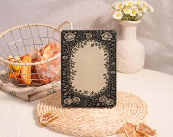 May include: A rectangular metal tin featuring a black and white floral border with detailed roses and vines surrounding a central blank oval area. The tin is positioned upright on a round woven mat, showcasing a high-contrast botanical design.