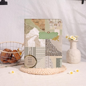 May include: A rectangular journal featuring a collage-style cover with sage green, beige, and cream tones. The design includes a gold-toned circular frame with the text "to the moon and back, remember?" and a green label with a yellow dragonfly graphic.