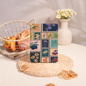 May include: A rectangular journal with a botanical stamp design. The cover features various floral illustrations in a grid pattern, including flowers, berries, and leaves, set against a backdrop of muted colors. The journal is placed on a woven mat.