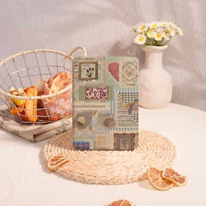 May include: A decorative journal with a patchwork design featuring floral, heart, and gingham patterns. The journal is on a woven placemat, with a basket of bread and a vase of flowers in the background.