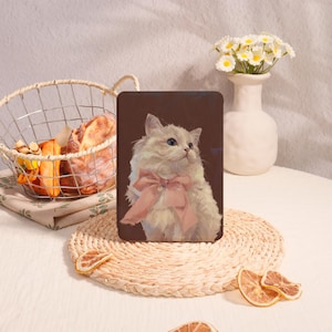 Op de afbeelding: Een tablethoes met een pluizige witte kat met blauwe ogen en een roze strik. De kat staat tegen een bruine achtergrond. De hoes ligt op een geweven placemat, met een broodmand en een vaas met bloemen op de achtergrond.