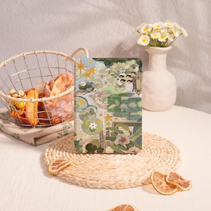May include: A decorative journal with a collage design featuring celestial elements, flowers, and insects. The cover has a green and beige color scheme with gold accents. The journal is placed on a woven placemat with dried orange slices.