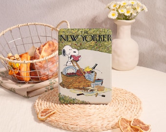 May include: A rectangular metal tin featuring a vintage The New Yorker magazine cover illustration of Snoopy and Woodstock having a picnic. The artwork depicts a picnic basket, food, and drinks on a blanket. The tin has a cream-coloured background.